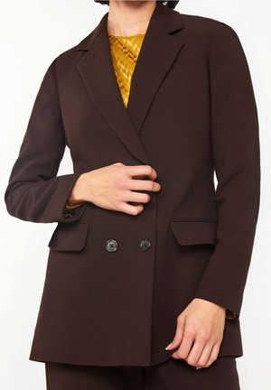 Persoon die een donkerbruin colbert aanpast over een mosterdgele top met patroon, met zicht op de voorste knopen en klepzakken, geknipt boven de nek.