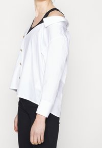 Personne portant une chemise blanche boutonnée tombée sur une épaule, laissant apparaître un débardeur noir en dessous, associée à un pantalon noir sur un fond uni.