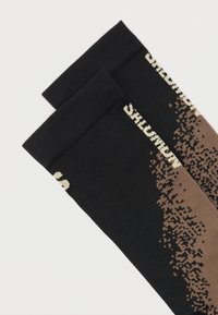 Ζευγάρι μαύρες κάλτσες με μπεζ pixel μοτίβο και λευκό κείμενο στο μανσέτα, παρουσιασμένες σε λευκό φόντο.