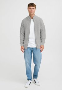 Grå strikket zip-up genser med høy krage, brukt over en hvit t-skjorte, paret med lyseblå jeans og hvite joggesko.