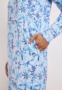 Abito a maniche lunghe in tessuto azzurro con un intricato motivo floreale nelle tonalità di blu. Include una tasca laterale e polsini aderenti.