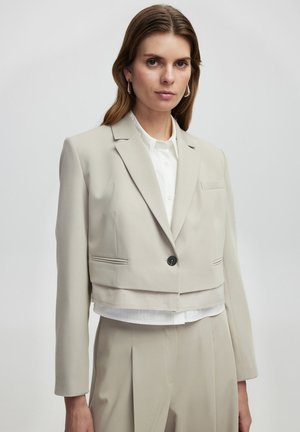 Korte lichtbeige blazer met een enkele zwarte knop, met een reverskraag en een voorzak; gedragen over een wit overhemd met kraag.