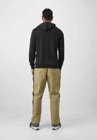 Sweatshirt noir à capuche avec des manches longues, associé à un pantalon beige clair avec des bandes latérales noires. Les chaussures sont grises avec une semelle blanche.
