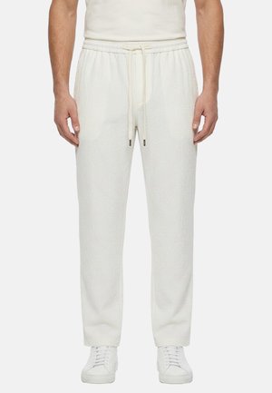 Uomo che indossa pantaloni bianchi con trama e vita con coulisse e sneakers bianche, in piedi davanti a uno sfondo semplice.