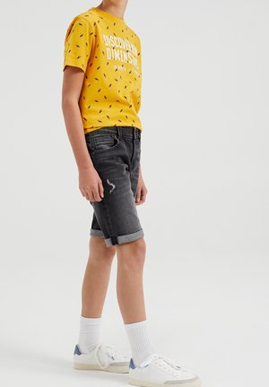 Żółta koszulka z czarnymi wzorami i białym tekstem, w połączeniu z czarnymi szortami jeansowymi z podwiniętymi mankietami i przetartym detalem. Białe trampki.