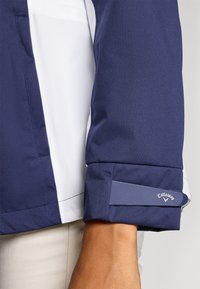 Tumesinine ja valge jakk, millel on sile tekstuur, kena disain, kontrastõmblused ja "Callaway" logo aksent varrukal.