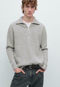 Giovane uomo che indossa un maglione grigio a mezza zip sopra una camicia bianca, con jeans scuri e una cintura in pelle intrecciata, mani in tasca.