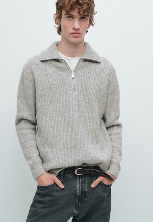 Junger Mann, der einen grauen Strick-Halbzippullover über einem weißen Hemd, dunklen Jeans und einem geflochtenen Ledergürtel trägt, mit den Händen in den Taschen.