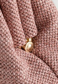 Záber zblízka na ružovú tkanú látku s textúrou a lesklým zlatým gombíkom, ktorý je čiastočne viditeľný pod záhybmi.