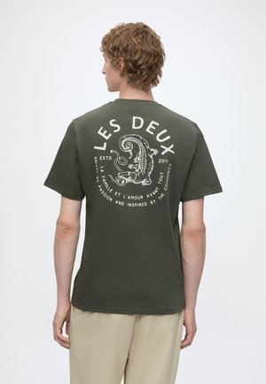 Personne aux cheveux bouclés portant un t-shirt vert olive avec un graphisme crocodile blanc et un texte en français imprimé dans le dos.