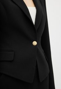 Blazer nero realizzato in tessuto strutturato, con rever a punta, due tasche frontali e un bottone rotondo dorato in vita.