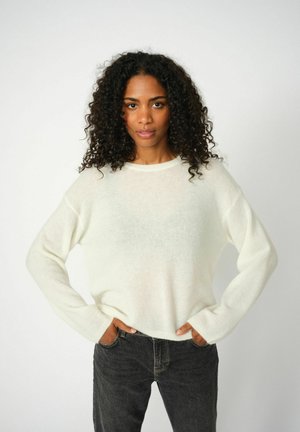 Vrouw met krullend haar in een witte trui en zwarte spijkerbroek staat met haar handen in de zakken tegen een effen lichte achtergrond.