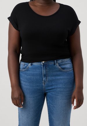 Person wearing a black short-sleeve top with lace trim and blue denim jeans, standing against a plain background.