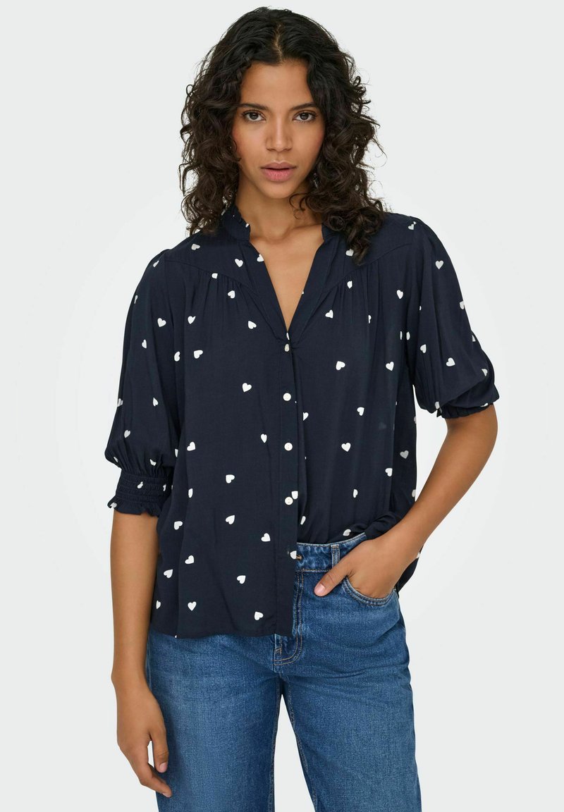 Marinebluse med hvidt hjerte mønster, knaplukning foran, og korte puffærmer. Lavet af letvægtsstof med en afslappet pasform.