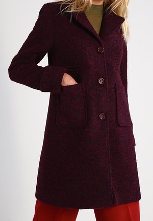 Femme portant un manteau en laine bordeaux foncé avec de gros boutons et des poches plaquées, associé à un pantalon rouge et un haut vert olive.