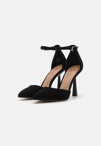 Paire d'escarpins noirs à bout pointu avec brides à la cheville et boucle dorée, semelle intérieure beige, sur fond blanc.