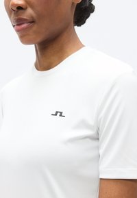 Άσπρη κοντομάνικη μπλούζα από λείο ύφασμα, με μικρό μαύρο λογότυπο στην αριστερή πλευρά του στήθους. Απλός στρογγυλός γιακάς, άψογη ραφή χωρίς ραφές.