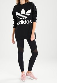 Černá mikina s velkým bílým logem Adidas, v kombinaci s černými legínami s síťovanými panely. Dokončeno s růžovými texturovanými sportovními botami.