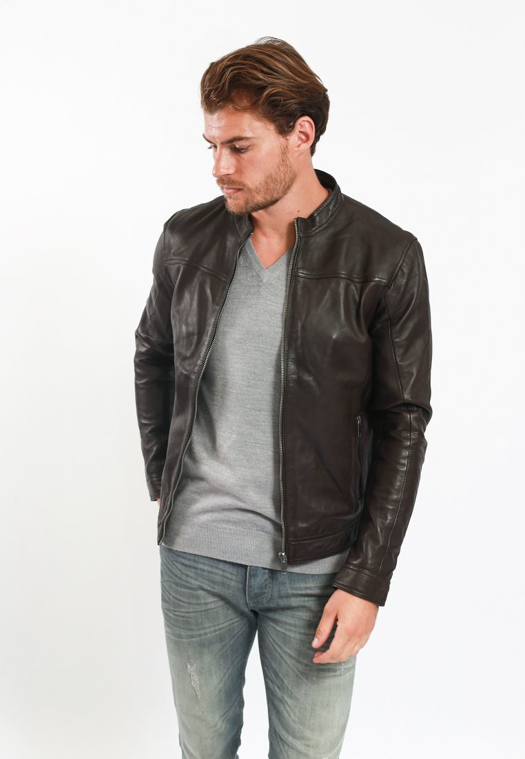 Giacca in pelle marrone scuro con design aderente, chiusura con zip e dettagli minimalisti. Indossata sopra un maglione a collo a V grigio chiaro e jeans chiari.