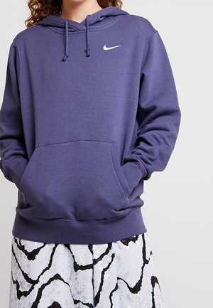 Persoon draagt een paarse Nike-hoodie met handen in de voorzak en een witte rok met zwarte abstracte patronen.