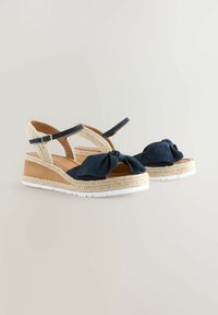 Kenkäplat ovat tummansinistä mokkaa, niissä on solmittu etuosa ja beige espadrillityylinen korko. Säädettävä nilkkahihna kultaisella soljella.