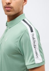 Homme portant un polo vert clair avec un détail d'épaule à rayures blanches et noires et le texte "Peak Performance" sur la manche.