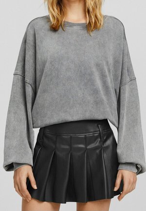 Kobieta w oversize szarej bluzie z długim rękawem i czarnej plisowanej skórzanej mini spódniczce, stoi z rękami swobodnie opuszczonymi wzdłuż ciała.