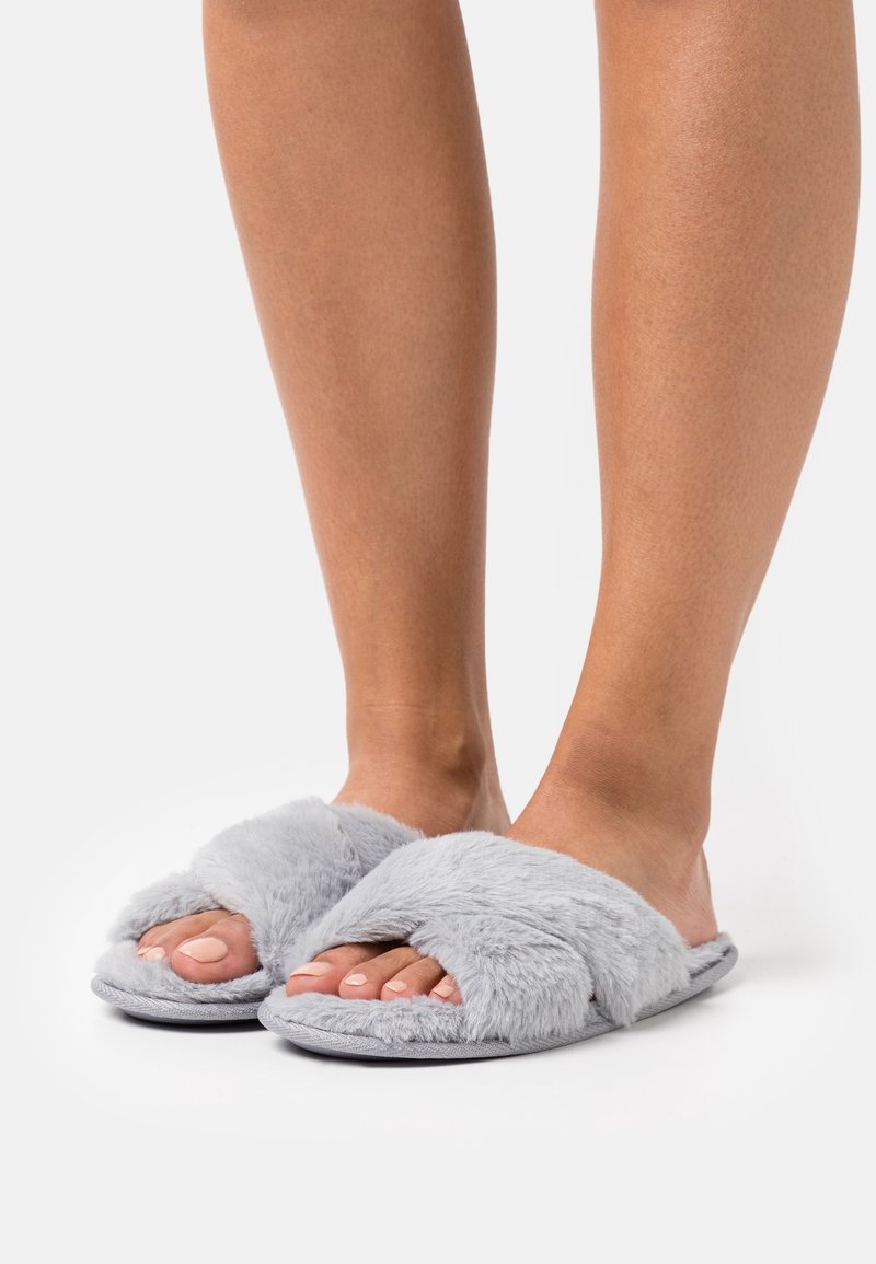 Pieds portant des chaussons gris pelucheux à bouts ouverts avec des brides croisées sur une surface blanche.