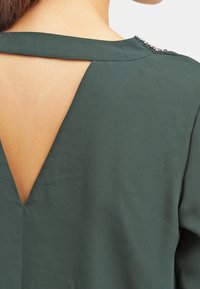 Grøn stoftop med V-udskæring i ryggen. Har metallisk accent nær udskæringen. Glat tekstur, struktureret design.