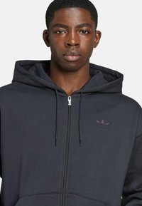 Black zip-up hoodie made of cotton blend fabric. Features a kangaroo pocket, ribbed cuffs, and a subtle embroidered logo on the chest.