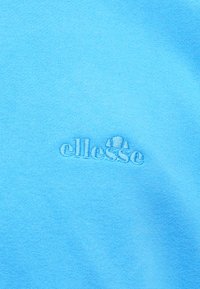 Plava tkanina s malim, izvezenim ellesse logotipom koji prikazuje stiliziranu polovicu teniske lopte i skijaške vrhove u pratećoj plavoj niti.