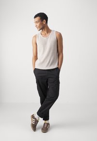 Világosszürke bordázott ujjatlan felső, fekete cargo nadrág oldalsó zsebekkel és barna sneakers fehér csíkokkal. A modell a kezeit a zsebében tartja.