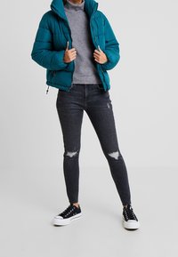 Teal pufferjack, hoge kraag, ritssluiting en verticale naden; grijze coltrui, donkere gescheurde skinny jeans en zwarte sneakers.