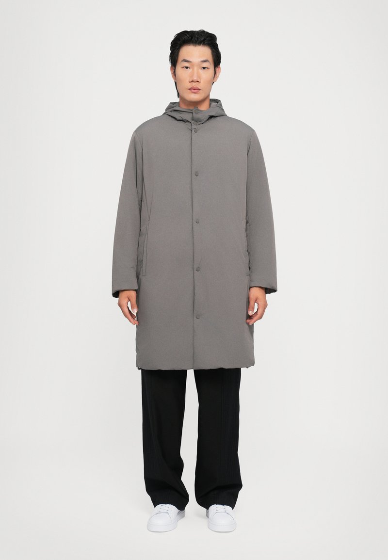 Manteau gris rembourré avec capuche, boutons snap à l'avant et poches latérales, associé à un pantalon noir et des baskets blanches. Design simple et fonctionnel.