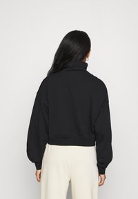 Sweatshirt noir raccourci avec un col montant, des manches longues et des poignets côtelés. Le matériau semble doux et texturé, conçu pour une coupe décontractée.