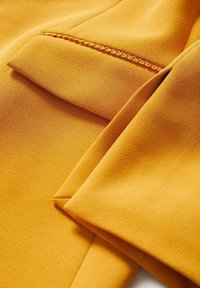 Tissu jaune moutarde avec un gros plan sur un rabat de poche et un détail de couture ornementale en boucle.