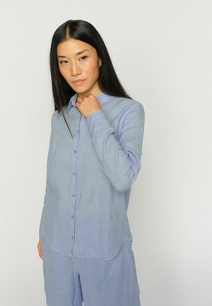 Žena s dlhými čiernymi vlasmi, ktorá má oblečenú svetlomodrú košeľu na gombíky a zodpovedajúce šortky, drží si golier jednou rukou.