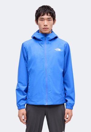 Młody mężczyzna noszący jasnoniebieską bluzę z kapturem The North Face z suwakiem z przodu i czarne spodnie, stojący na jednolitym tle.