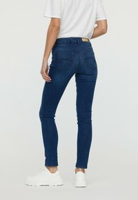 Jean skinny bleu foncé taille haute, avec deux poches arrière et des détails de couture, associés à des baskets blanches et une chemise à manches courtes.