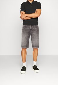 Mies, joka käyttää mustaa polo-paitaa, haalistuneita harmaita farkkushortseja, valkoisia sukkia ja mustia lenkkikenkiä, seisoo kädet ristissä.