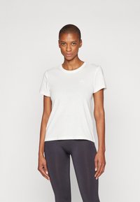 White cotton t-shirt with a crew neck, short sleeves, and a subtle logo detail on the chest, paired with dark leggings.