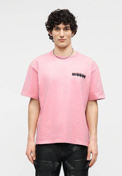 Giovane con capelli ricci che indossa una maglietta rosa a maniche corte con scritta nera sul petto, pantaloni cargo neri e una collana d'argento.