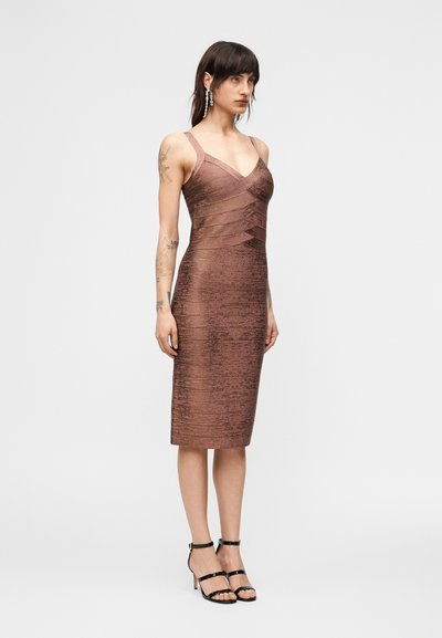 Γυναίκα που στέκεται φορώντας εφαρμοστό, αμάνικο, υφασμένο μπρούτζινο μίντι φόρεμα και μαύρα ψηλοτάκουνα σανδάλια με λουράκια, με τατουάζ στα χέρια και κρεμαστά σκουλαρίκια.