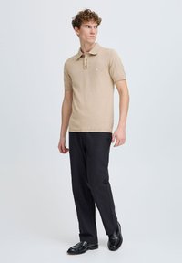 Polo bej cu mânecă scurtă, cu guler, având un sistem de închidere cu trei nasturi și manșete ribbate, asortat cu pantaloni negri și pantofi negri lucioși.