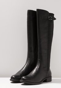 Bottes noires montantes en cuir lisse avec un panneau en textile tissé à l'arrière, présentant un petit talon et un accent de boucle décoratif.