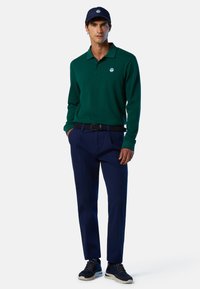 Polo verde de manga larga con cuello y logo. Combinado con pantalones azul marino y zapatillas negras. El cinturón es de tela tejida.