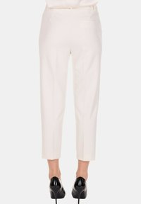 Gambe che indossano pantaloni bianchi su misura a vita alta e scarpe col tacco nere, viste da dietro su uno sfondo semplice.