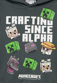 Σκούρο φούτερ με κουκούλα με λευκή πλακέ κείμενο «Crafting Since Alpha», περιβαλλόμενο από pixelated κεφάλια χαρακτήρων του Minecraft και το λογότυπο του Minecraft από κάτω.