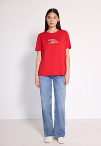 Camiseta de algodón roja con mangas cortas y cuello redondo, que presenta un texto blanco que dice "evoluciona, no compitas". Combinada con unos jeans acampanados de color azul claro.
