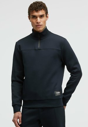 Dunkelblaues Half-Zip Sweatshirt mit erhöhtem Kragen, flachen Nähten und einem Logo-Patch auf der unteren linken Seite. Weicher Stoff, schlanker Schnitt, minimalistisches Design.